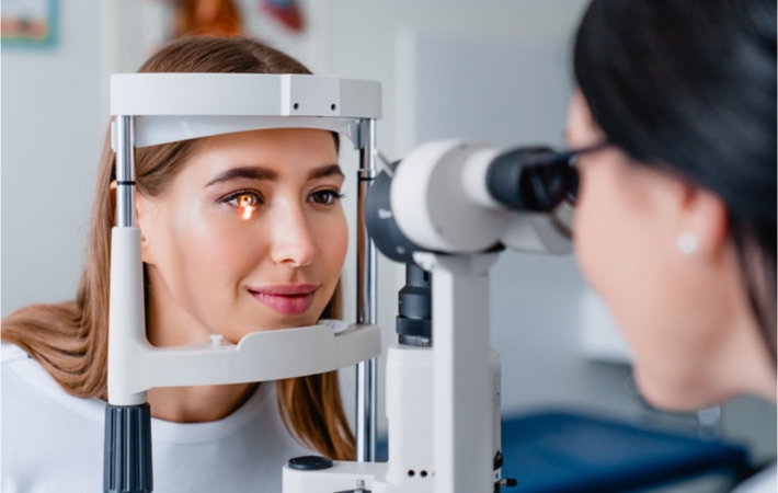 A female optometrist examines a young smiling brunette patient with a Slit Lamp.