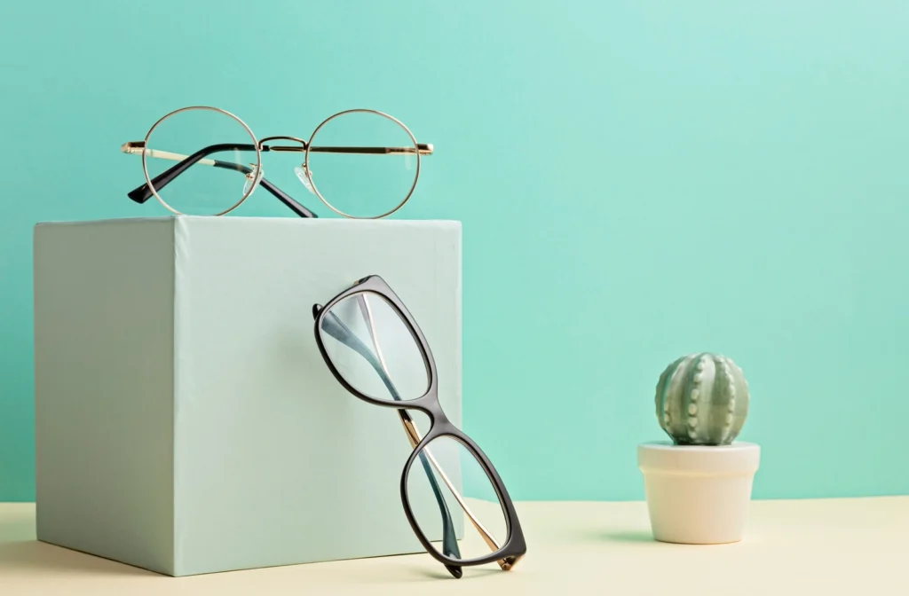 Against a lime green background a lightly shaded green design box lain upon a shelf has a pair of glasses sitting on top, with a second pair of glasses leaning against it.