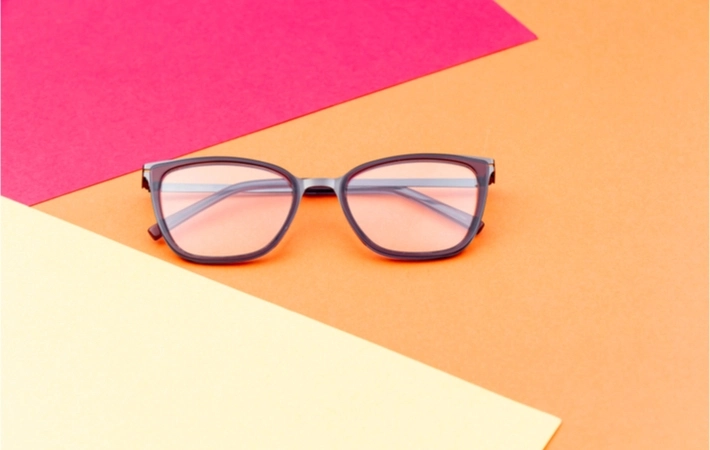 A pair of black glasses is laid on top of a mixed colour background with orange, pink, and yellow.