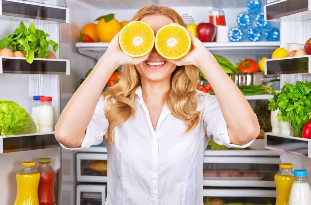 A person holds 2 orange halves in front of her eyes while standing before a fridge full of healthy foods