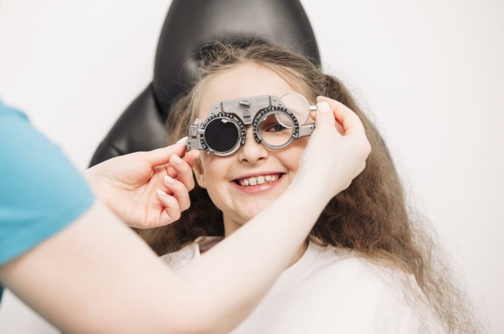 Little girl with low brown pigtails smiling while peering through optometrist vision device. Doctor's hands are pictured in frame holding the device up to the child's face.