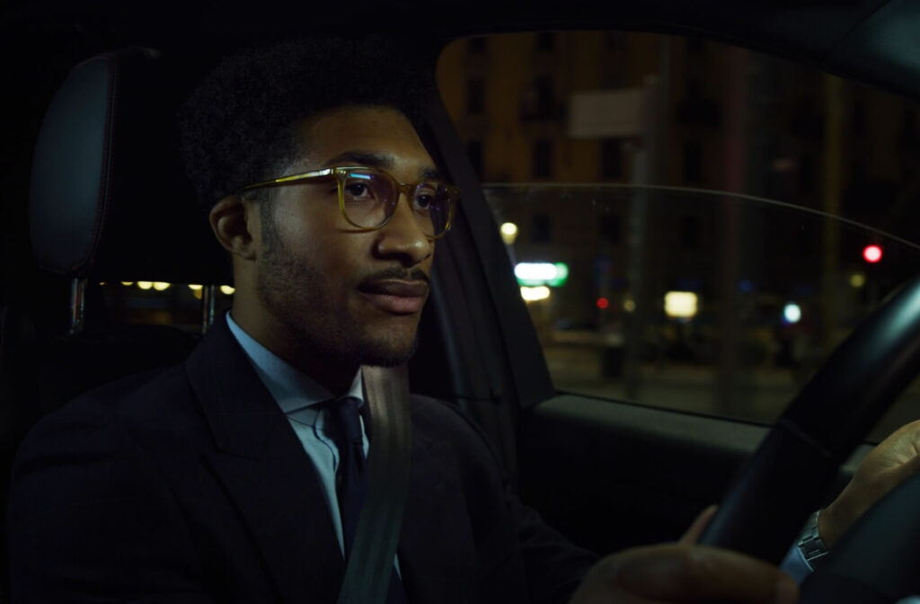 A driver wearing glasses and a seatbelt, focused on the road while driving at night in an urban area.