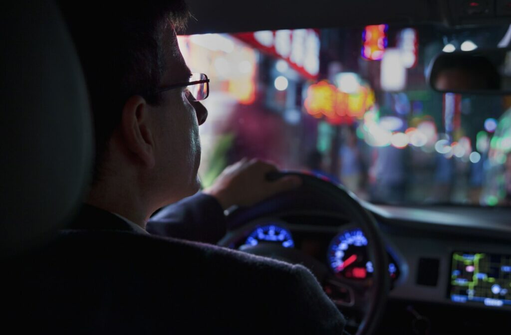 A person with glasses driving at night with neon city lights reflecting through the car windshield.