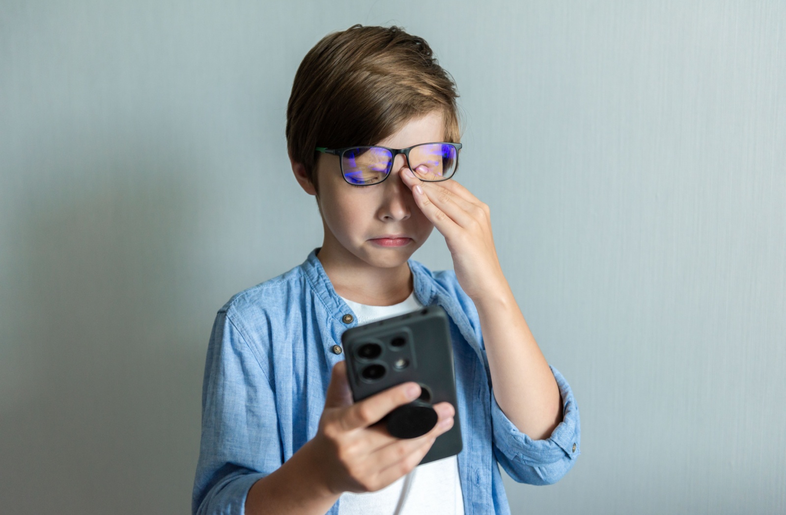 A child wearing glasses, rubbing his eyes while looking at a smartphone