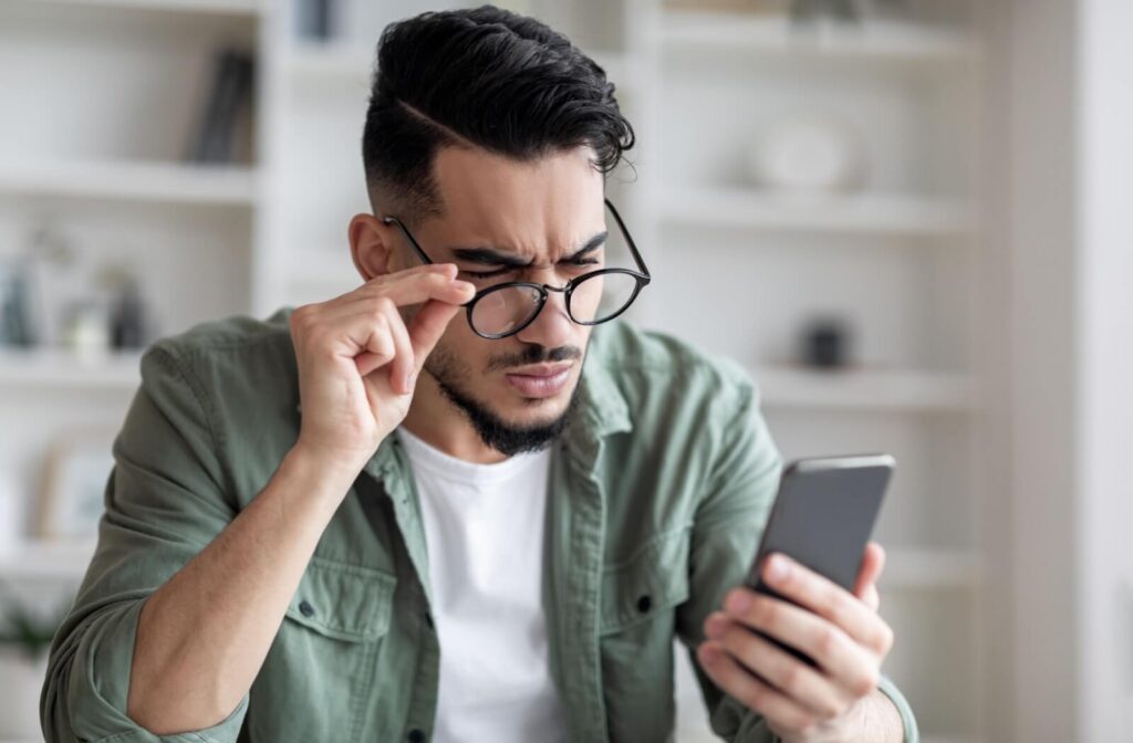 A man squinting and adjusting his eyeglasses while looking at his smartphone, illustrating blurry vision from astigmatism.