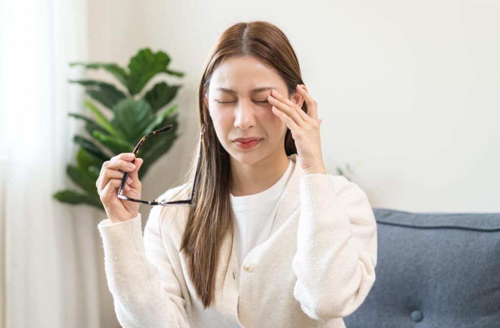 A woman sitting on a couch rubbing her closed, irritated eye while holding her tortoiseshell glasses, illustrating symptoms of itchy allergy eyes or pink eye.