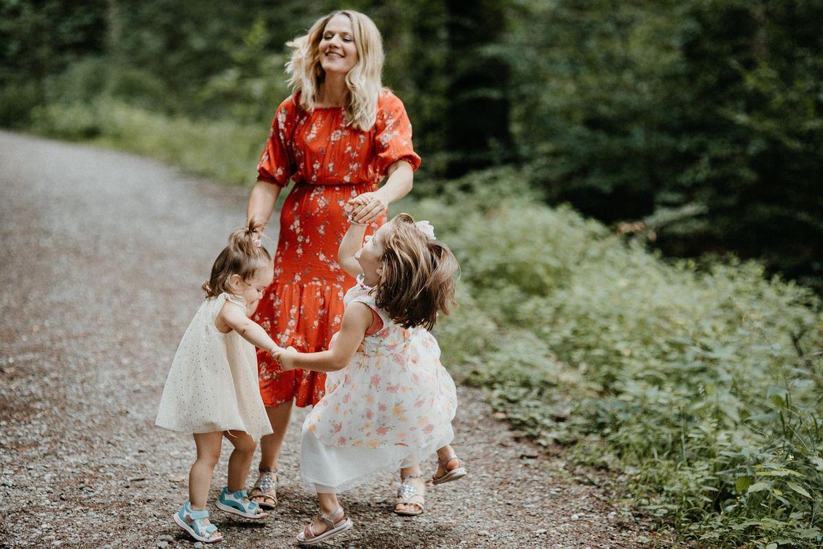 Smiling adult in a red floral dress watches two young children in dresses hold hands and spin on a gravel path surrounded by green trees.